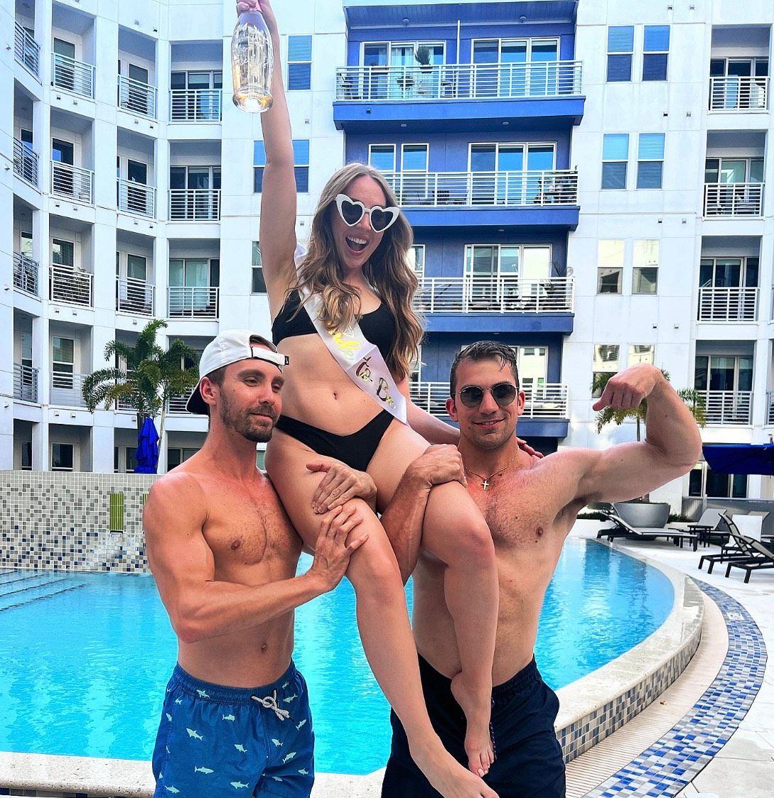 Woman in black bikini with heart-shaped sunglasses and a sash holding a bottle, lifted by two shirtless men at a modern hotel pool with white-and-blue balconies