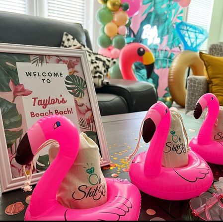 Tropical-themed indoor bachelorette party setup in a living room with a framed welcome sign, pink inflatable flamingo cup holders holding small favor bags, pastel balloon garland and palm-print backdrop on a table.