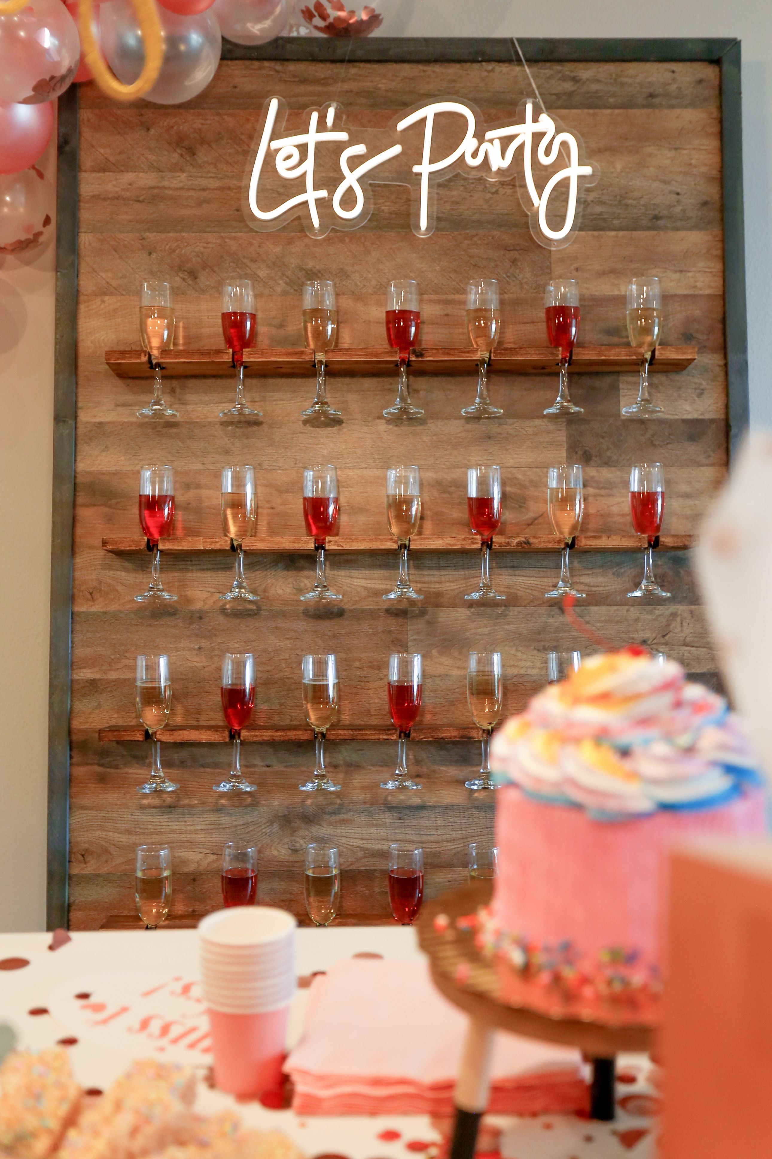 Festive party display: wooden champagne wall holding rows of wine flutes with rosé and sparkling wine beneath a glowing “Let’s Party” neon sign, with a pink frosted cake, stacked cups, pastel balloons and confetti on the table in the foreground.