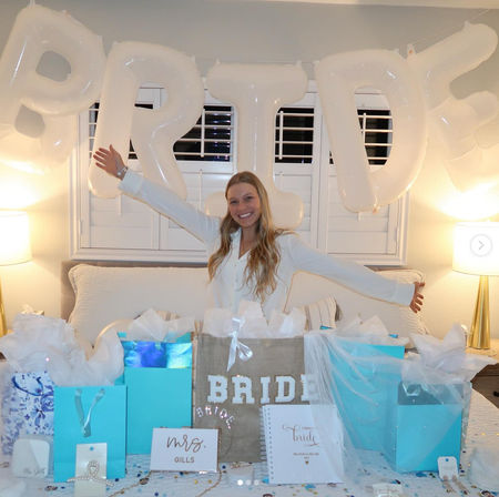 Beaming bride-to-be with arms outstretched on a bed beneath large white "BRIDE" balloon letters, surrounded by turquoise and white gift bags and presents in a cozy at-home bridal shower setting.
