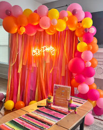 Indoor living room party setup with vibrant pink, orange and yellow balloon arch and streamers, glowing neon sign reading Let's Party, festive striped table runner and mini piñata decorations.