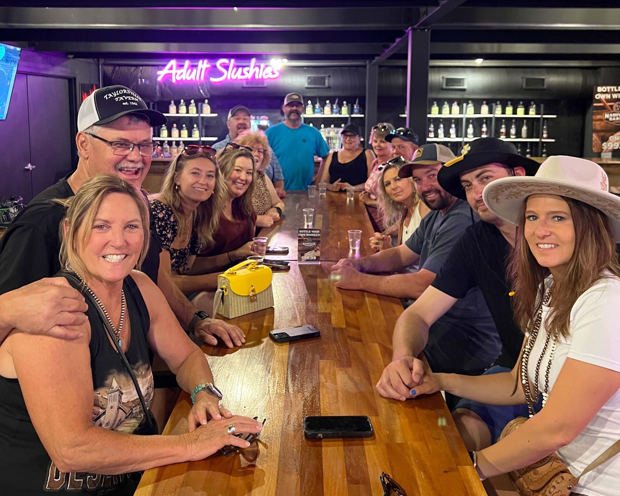 Smiling group of adults gathered around a long wooden bar enjoying drinks, with a neon “Adult Slushies” sign and backlit bottle shelves in a lively bar interior.