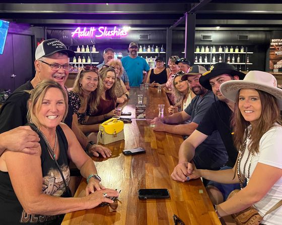 Smiling group of adults gathered around a long wooden bar enjoying drinks, with a neon “Adult Slushies” sign and backlit bottle shelves in a lively bar interior.