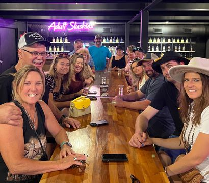 Smiling group of adults gathered around a long wooden bar enjoying drinks, with a neon “Adult Slushies” sign and backlit bottle shelves in a lively bar interior.