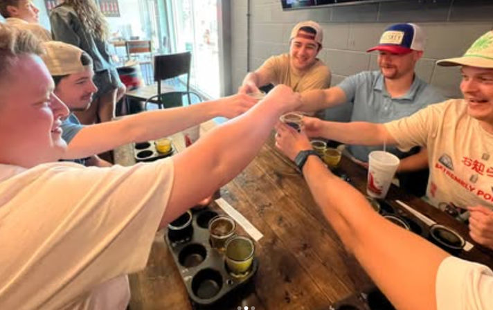 Group of friends clinking glasses and cheering over beer flight trays on a rustic wooden table at a casual bar.