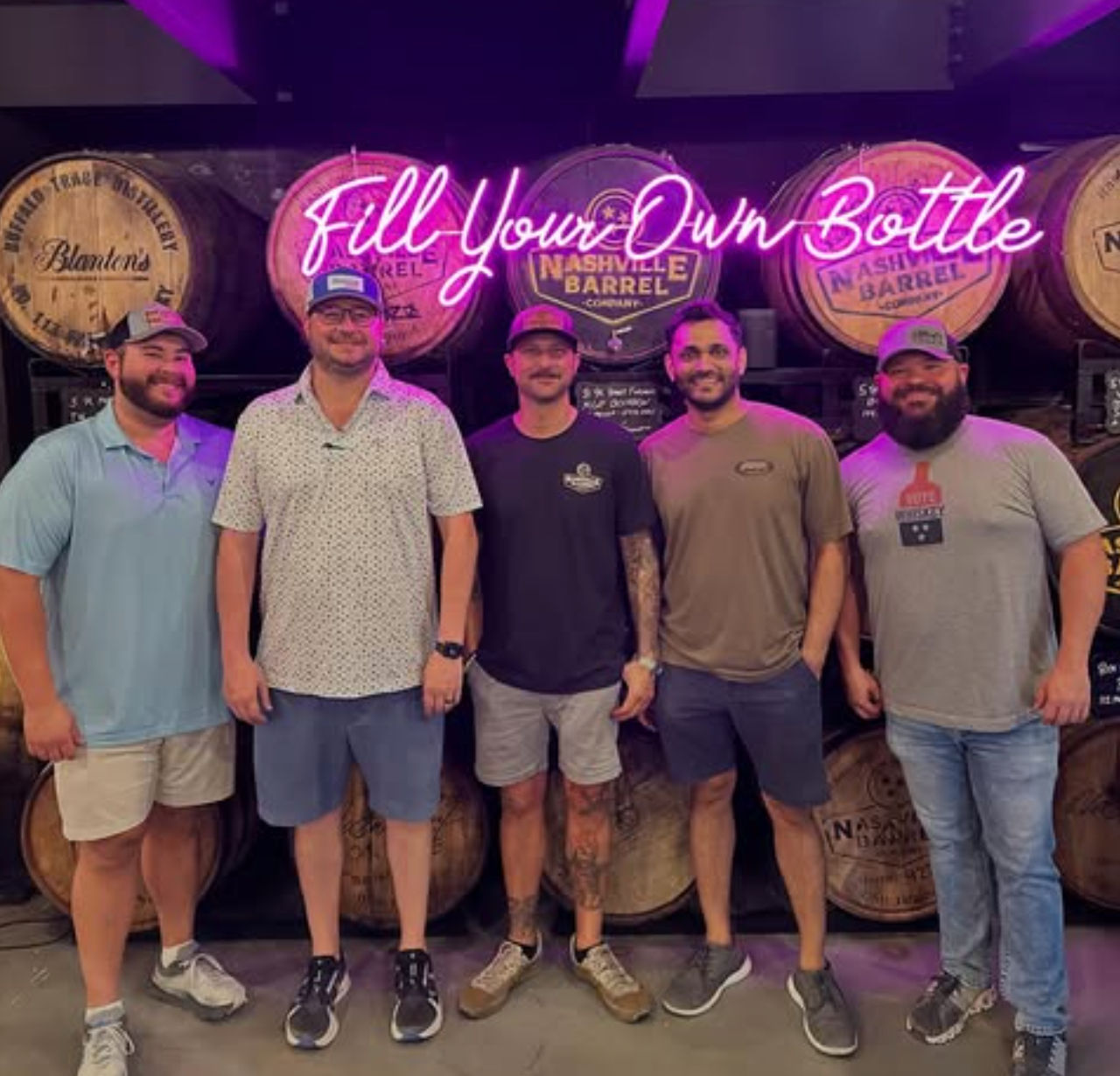 Five men smiling and posing in a Nashville distillery tasting room in front of stacked wooden whiskey barrels and a pink neon "Fill Your Own Bottle" sign.
