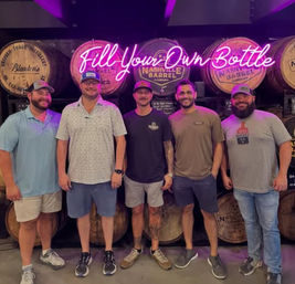 Five men smiling and posing in a Nashville distillery tasting room in front of stacked wooden whiskey barrels and a pink neon "Fill Your Own Bottle" sign.