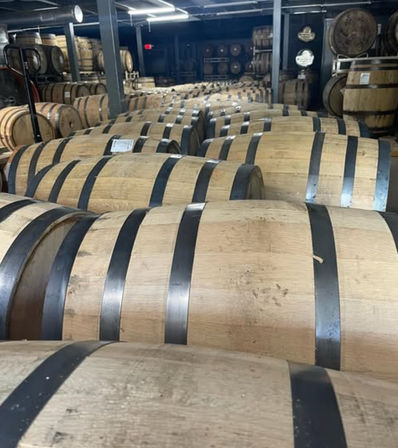 Rows of oak barrels with metal hoops in a dim distillery barrel room, stacked for aging whiskey and other spirits.