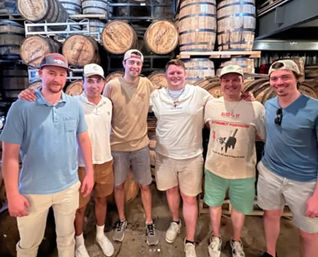 Six casually dressed men smiling and posing in a craft distillery barrel room in front of stacked oak whiskey barrels on racks.