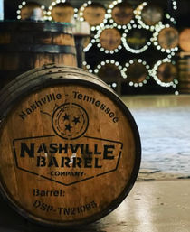 Rustic oak whiskey barrel stamped "Nashville, Tennessee" in foreground, stacked barrels and twinkling circular bokeh lights in a blurred distillery warehouse background.