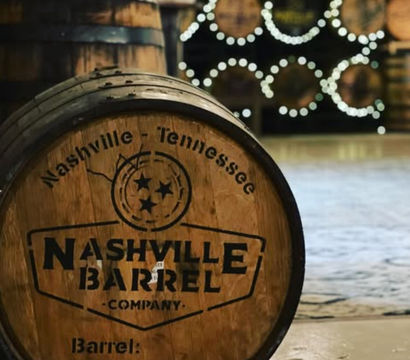 Rustic oak whiskey barrel stamped "Nashville, Tennessee" in foreground, stacked barrels and twinkling circular bokeh lights in a blurred distillery warehouse background.