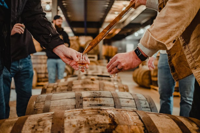 Hands using a whiskey thief to pour amber whiskey into tasting glasses over oak aging barrels in a craft distillery warehouse