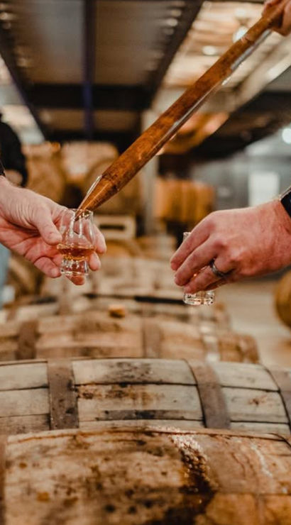 Hands using a whiskey thief to pour amber whiskey into tasting glasses over oak aging barrels in a craft distillery warehouse
