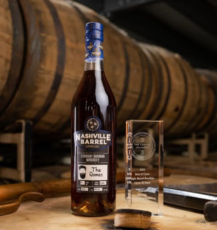 Dark amber bottle of small-batch Tennessee bourbon with a blue cap and custom label beside a clear glass award, set on a barrel head amid stacked oak aging barrels in a distillery.