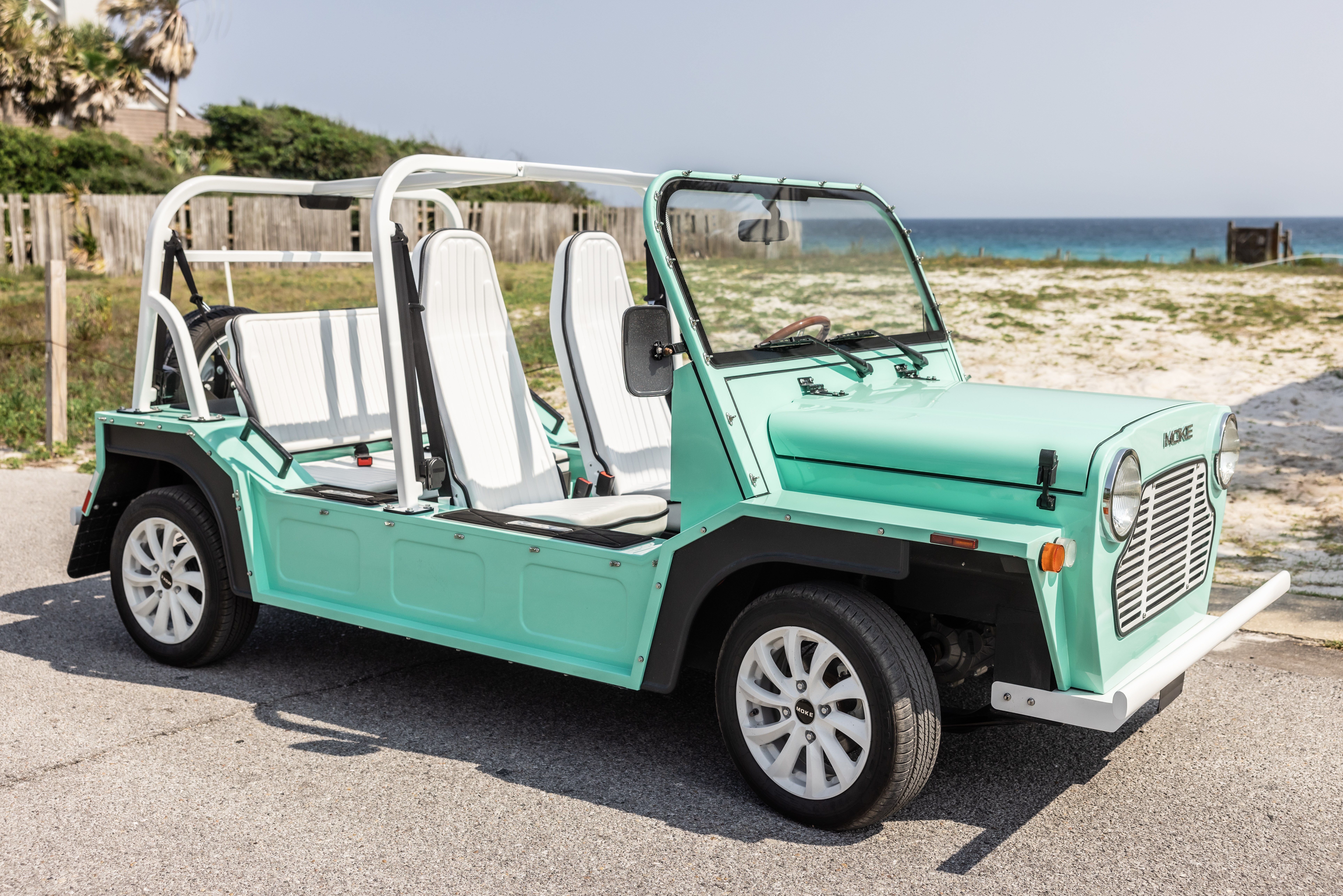 Retro mint-green open-top beach buggy with white seats parked on a seaside road, sand dunes and ocean in the background.