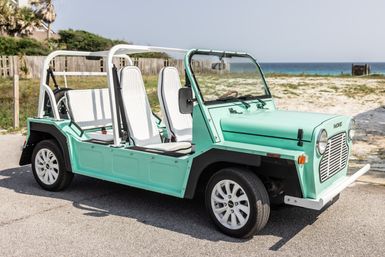 Retro mint-green open-top beach buggy with white seats parked on a seaside road, sand dunes and ocean in the background.