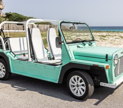 Retro mint-green open-top beach buggy with white seats parked on a seaside road, sand dunes and ocean in the background.