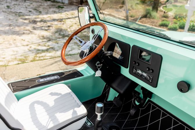 Sunlit mint-green vehicle interior with a polished wooden steering wheel, white quilted seat, black diamond-pattern floor mat, gear lever and dashboard radio controls, parked beside a sandy beach