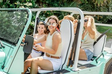 Four women in summer outfits smiling in a mint-green open-top beach buggy on a sunny drive, wearing sunglasses and casual shorts.