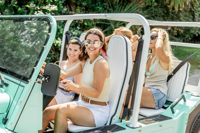 Four women in summer outfits smiling in a mint-green open-top beach buggy on a sunny drive, wearing sunglasses and casual shorts.