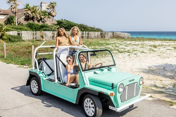 Four friends in a mint-green open-top beach car on a sunny coastal road beside sandy dunes and turquoise ocean, summer vacation vibe.