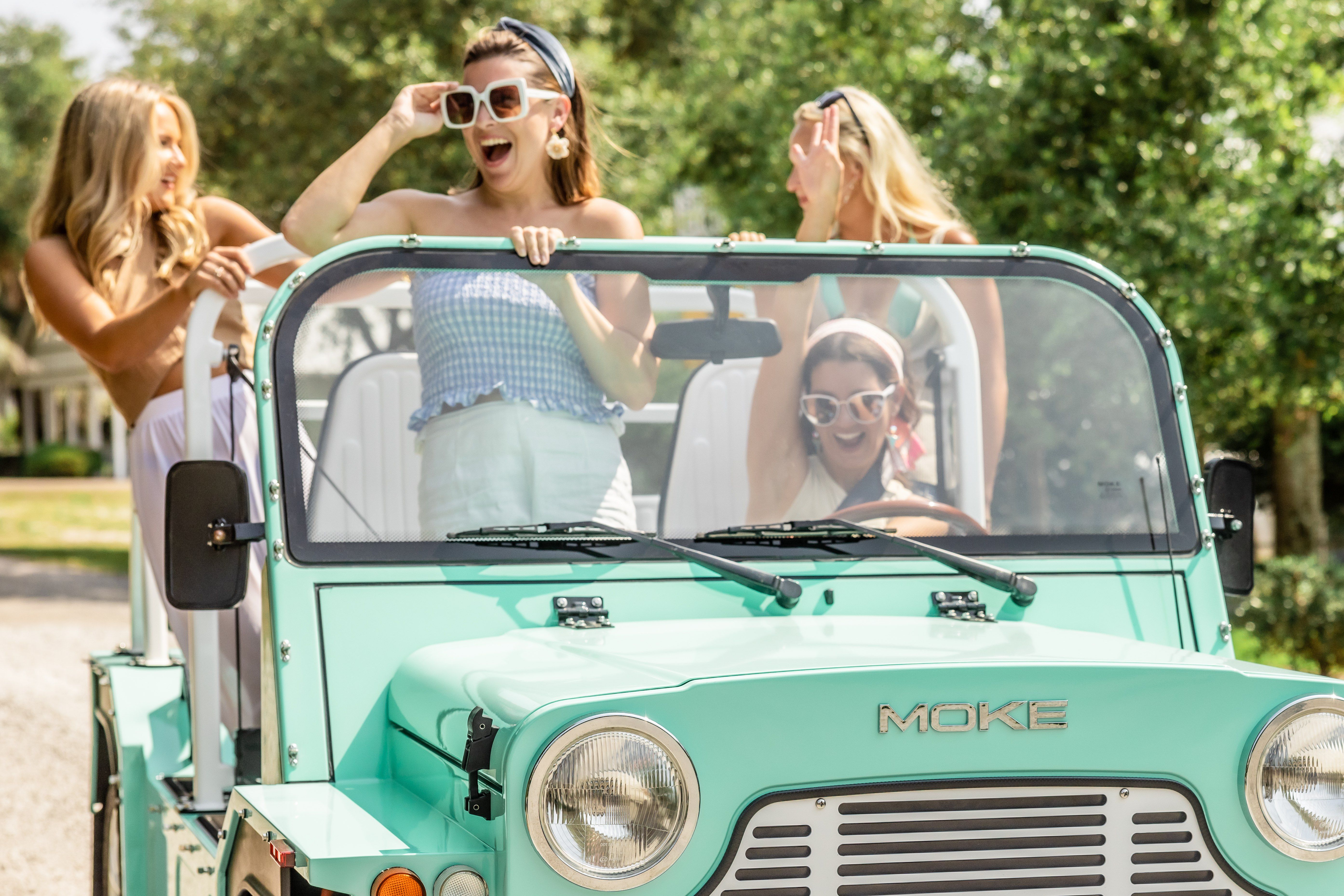 Four friends laughing in a mint-green vintage-style convertible on a sunny tree-lined street, wearing sunglasses and summer outfits.