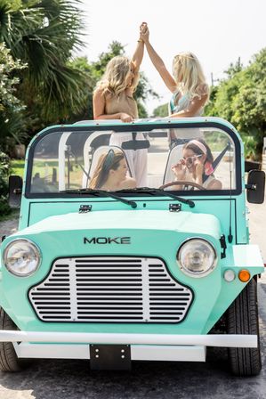 Four friends in a mint-green open-top beach car on a sunny coastal road, two standing and high-fiving while two sit and laugh.
