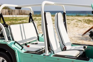 Mint-green open-top beach buggy with white ribbed seats parked on a sandy shore, blue ocean and dunes in the background