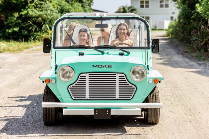 Front view of a mint-green open-top vintage beach car with four smiling women driving down a sunny gravel coastal lane, palm trees and a house in the background.