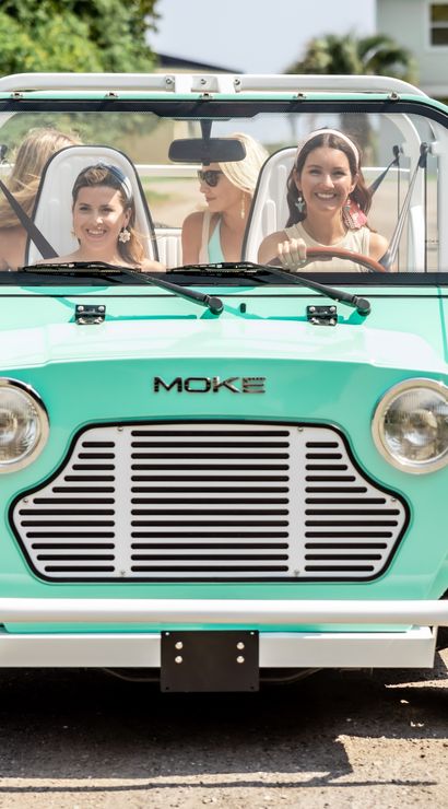 Front view of a mint-green open-top vintage beach car with four smiling women driving down a sunny gravel coastal lane, palm trees and a house in the background.