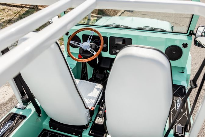 Top-down view of a sunlit mint-green open-top two-seat retro vehicle interior with white vinyl seats, wooden steering wheel, minimalist dashboard and black floor mats, parked outdoors.