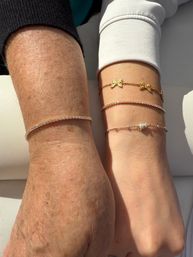 Close-up of two wrists side by side wearing stacked delicate bracelets — gold butterfly chain, slim diamond tennis bracelet, and a small pearl beaded chain on a sunlit white background.