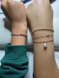Close-up of two forearms side by side showcasing wrist jewelry: left wrist with a silver chain bracelet featuring a small diamond-shaped link; right wrist styled with three layered gold-tone bracelets — a sparkly rhinestone tennis chain, a delicate chain with a tiny heart charm, and a thin chain with a white circular charm — fashion wrist accessory shot.