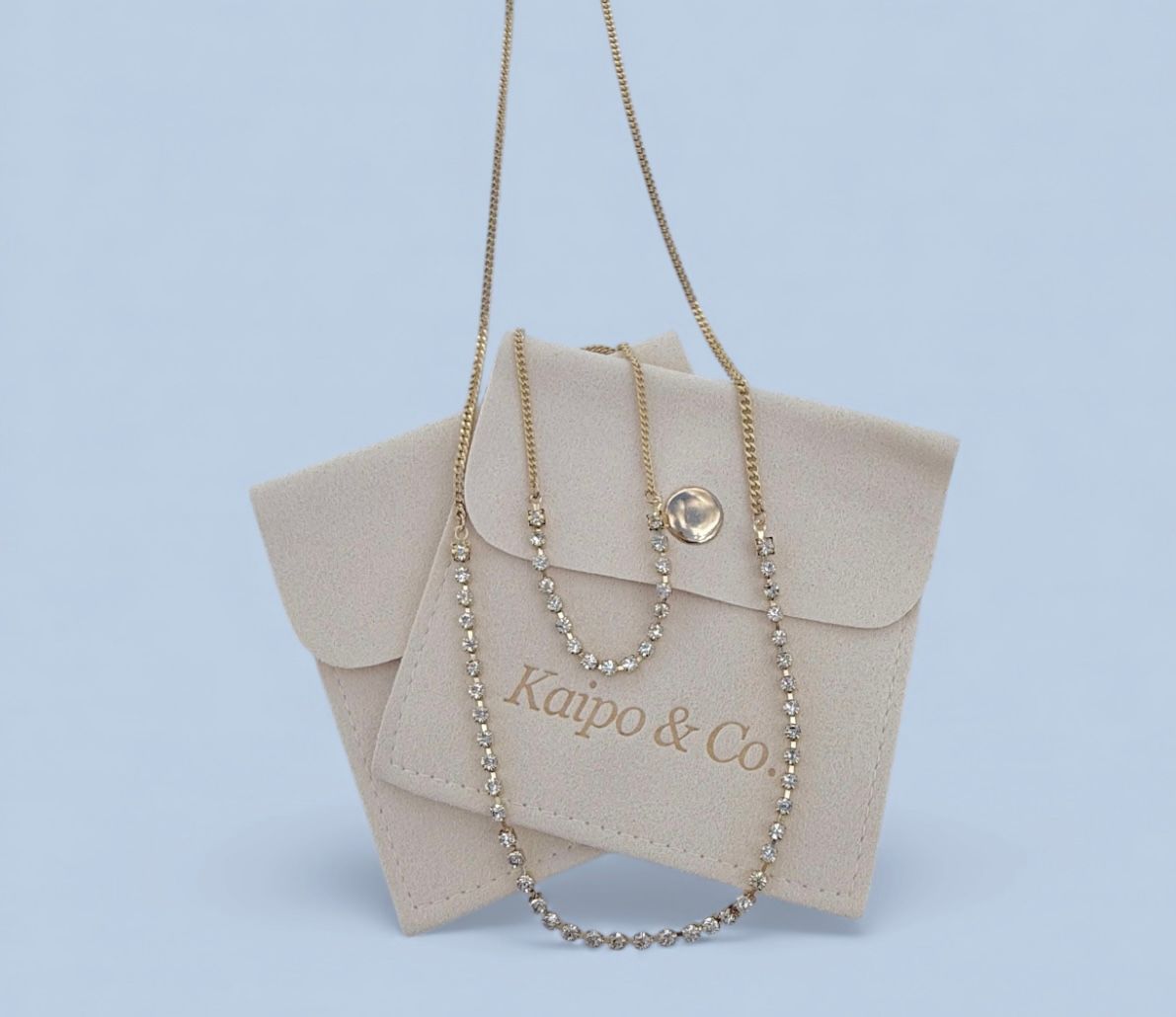 Two delicate gold necklaces — a fine chain and a sparkling rhinestone tennis strand — draped over a beige suede jewelry pouch on a pale blue background.