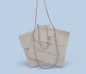Two delicate gold necklaces — a fine chain and a sparkling rhinestone tennis strand — draped over a beige suede jewelry pouch on a pale blue background.