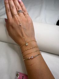Hand with light pink nails wearing layered delicate gold bracelets and a dainty hand-chain with small gemstone and pink-stone bangle on white fabric