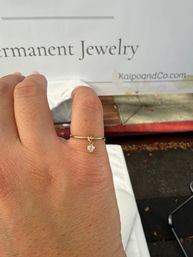Close-up of a hand wearing a thin gold ring with a dangling heart-shaped crystal charm, blurred outdoor jewelry stall sign in the background.