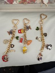 Flat-lay of three gold heart-clasp charm keychains on white fabric featuring colorful enamel charms — popcorn, fries, croissant, soda, playful cat and dog faces, leaves and novelty moon and mask pendants; fun accessory display, ideal for gifts or market booths.