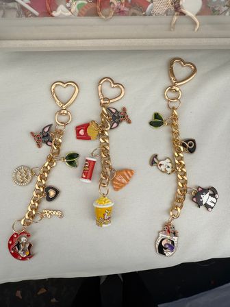 Flat-lay of three gold heart-clasp charm keychains on white fabric featuring colorful enamel charms — popcorn, fries, croissant, soda, playful cat and dog faces, leaves and novelty moon and mask pendants; fun accessory display, ideal for gifts or market booths.