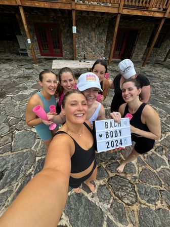 Group selfie of seven women in swimwear on a rustic stone patio outside a wooden lodge, holding pink cups and a lightbox sign reading "BACH BODY 2024"