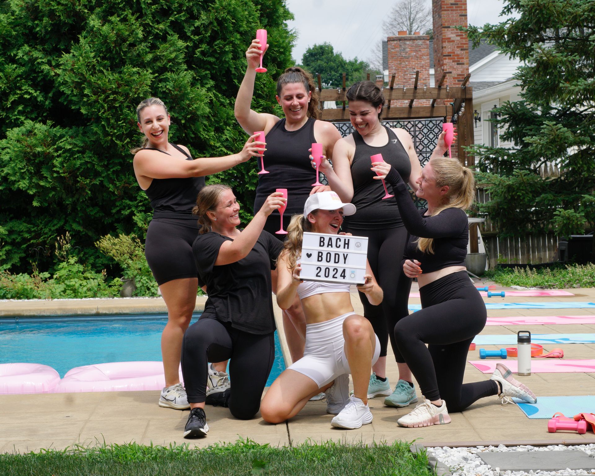 Six women in workout clothes toasting with pink flutes by a backyard pool, one kneeling in white holding a lightbox reading 'BACH BODY 2024' during an outdoor bachelorette-style fitness celebration.