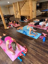 Group home-basement workout in a rustic finished space — women doing forearm planks on colorful yoga mats with pink dumbbells, resistance bands, a pool table and wooden staircase in the background.