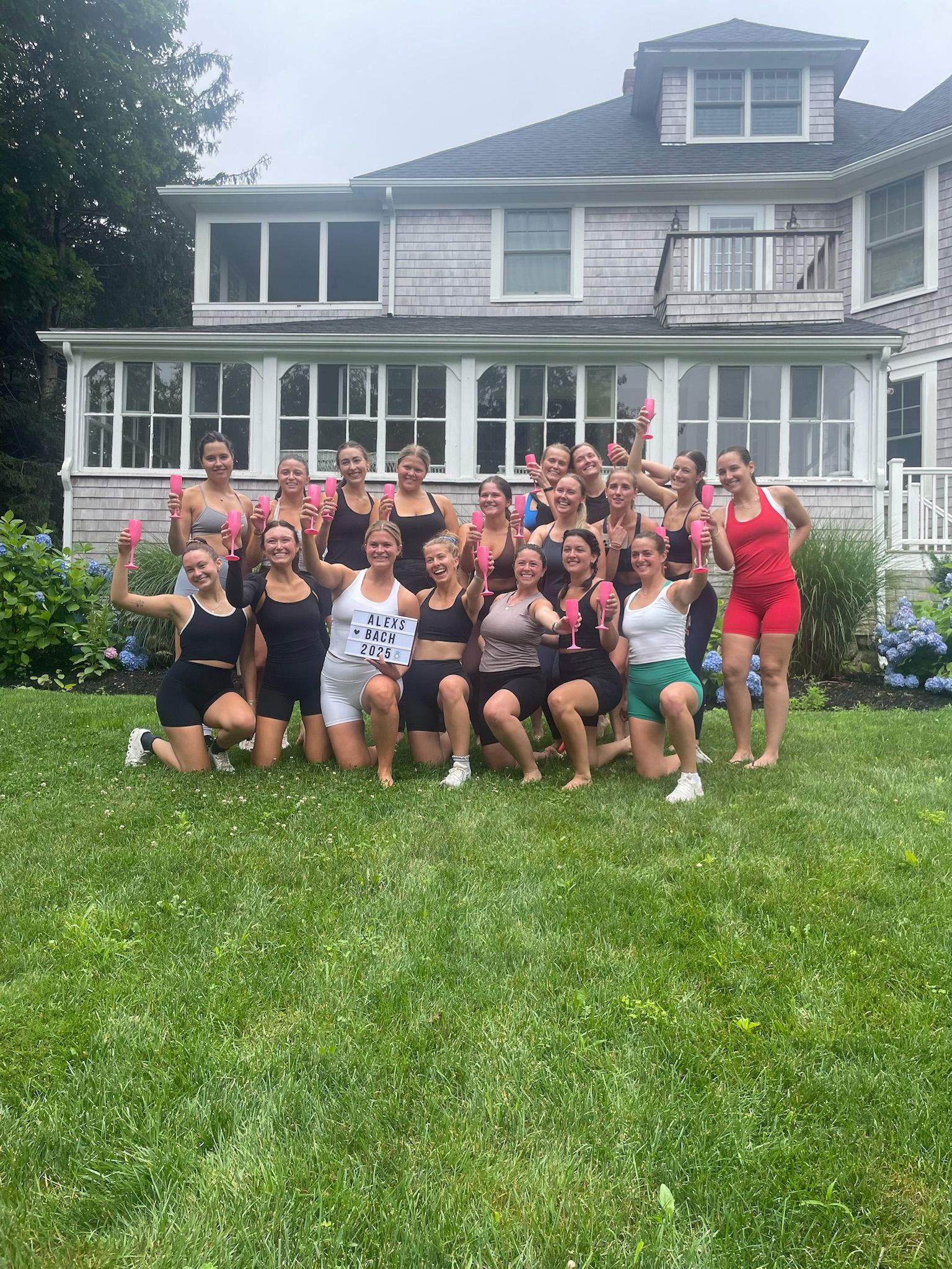 About 18 women in sporty summer outfits toasting with pink champagne flutes on a green backyard lawn in front of a shingle-sided coastal house, posing for a bachelorette celebration.
