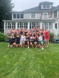 About 18 women in sporty summer outfits toasting with pink champagne flutes on a green backyard lawn in front of a shingle-sided coastal house, posing for a bachelorette celebration.