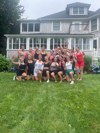 About 18 women in sporty summer outfits toasting with pink champagne flutes on a green backyard lawn in front of a shingle-sided coastal house, posing for a bachelorette celebration.