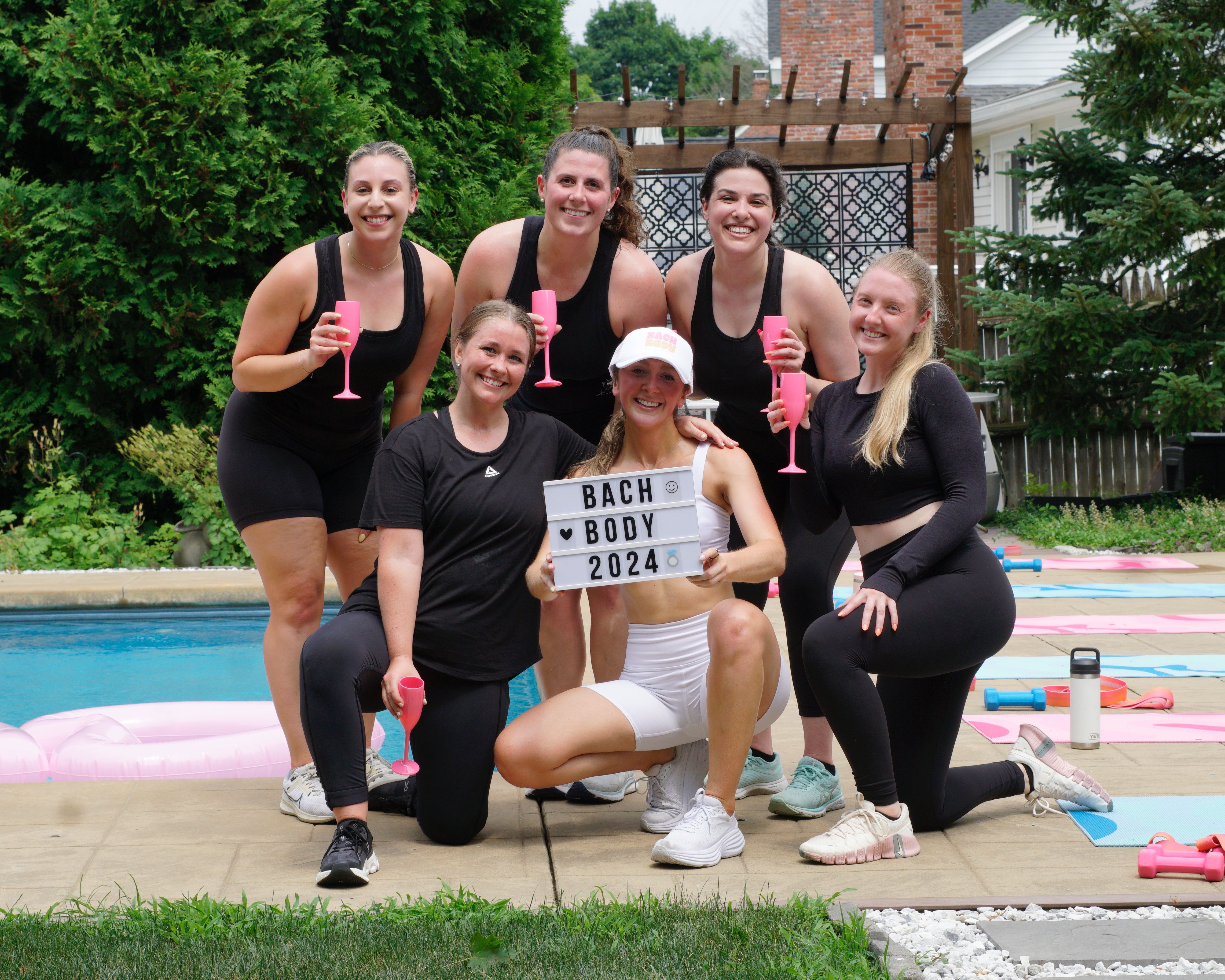 Six women in athletic wear posing poolside in a backyard, holding pink plastic flutes while the center woman kneels with a sign reading “BACH BODY 2024” surrounded by yoga mats and light dumbbells.