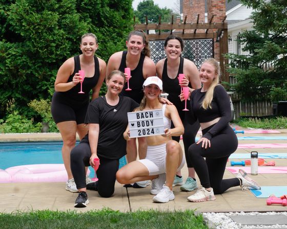Six women in athletic wear posing poolside in a backyard, holding pink plastic flutes while the center woman kneels with a sign reading “BACH BODY 2024” surrounded by yoga mats and light dumbbells.