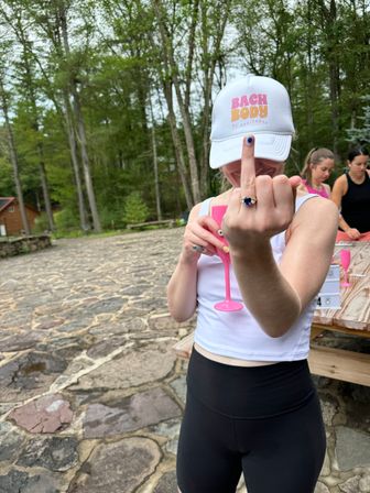 Woman at a wooded outdoor retreat on a stone patio, wearing a white cap and athletic outfit, holding a pink plastic champagne flute and playfully flipping the middle finger, picnic table and friends in the background.