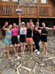 Six women in activewear cheering with pink champagne flutes on a stone patio in front of a rustic log cabin, holding a lightbox sign reading "Bach Body 2024".