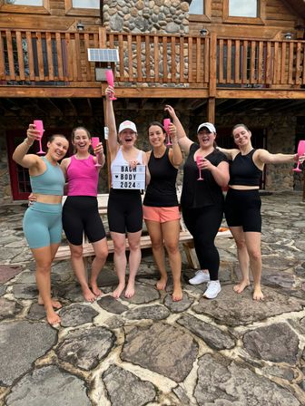 Six women in activewear cheering with pink champagne flutes on a stone patio in front of a rustic log cabin, holding a lightbox sign reading "Bach Body 2024".